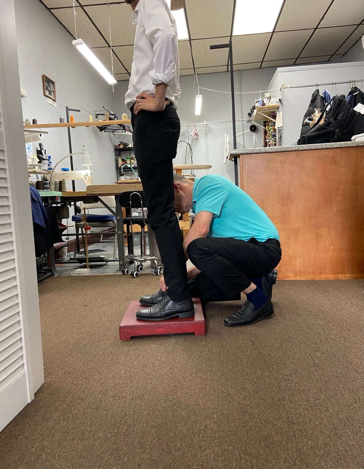 Nelson, the tailor, kneeling to pin the hem of a customer's trousers in the shop
